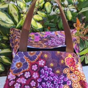 Sac cabas à motifs floraux colorés avec poignées marron, posé près de plantes vertes en extérieur.