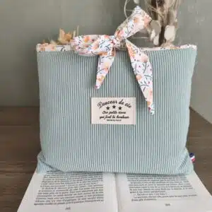 Pochette en tissu vert clair avec nœud floral derrière un livre ouvert sur une table en bois.