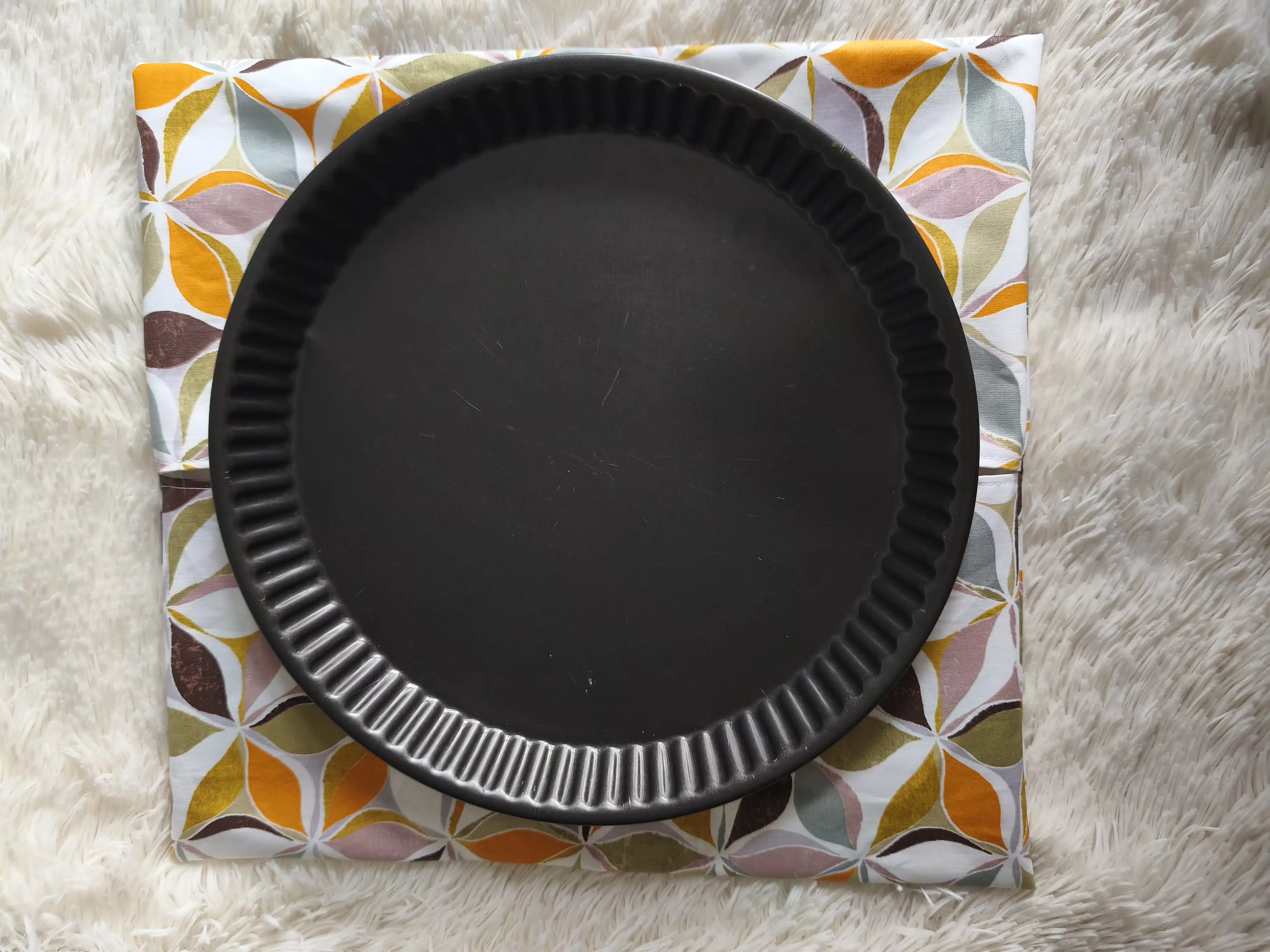 Sac à tarte 2 Moule à gâteau noir rond cannelé sur tissu à motifs, posé sur une surface blanche duveteuse.