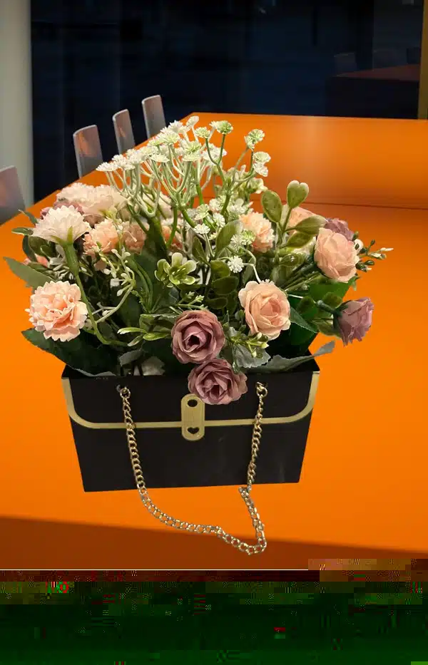 Sac à main noir à chaîne dorée avec fleurs artificielles sur table orange, chaises transparentes, décoration élégante.