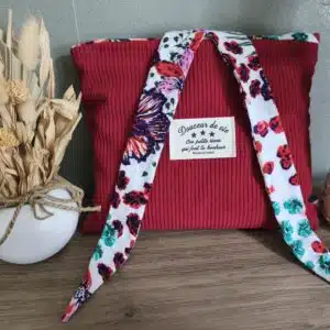 Pochette en tissu rouge avec lien fleuri, étiquette, sur bois, près de vases de fleurs séchées.