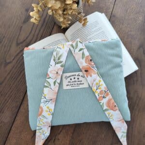 Pochette bleu clair en velours côtelé avec liens fleuris, sur table en bois près d'un livre ouvert et fleurs séchées.