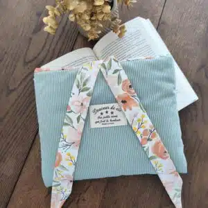 Pochette bleu clair en velours côtelé avec liens fleuris, sur table en bois près d'un livre ouvert et fleurs séchées.