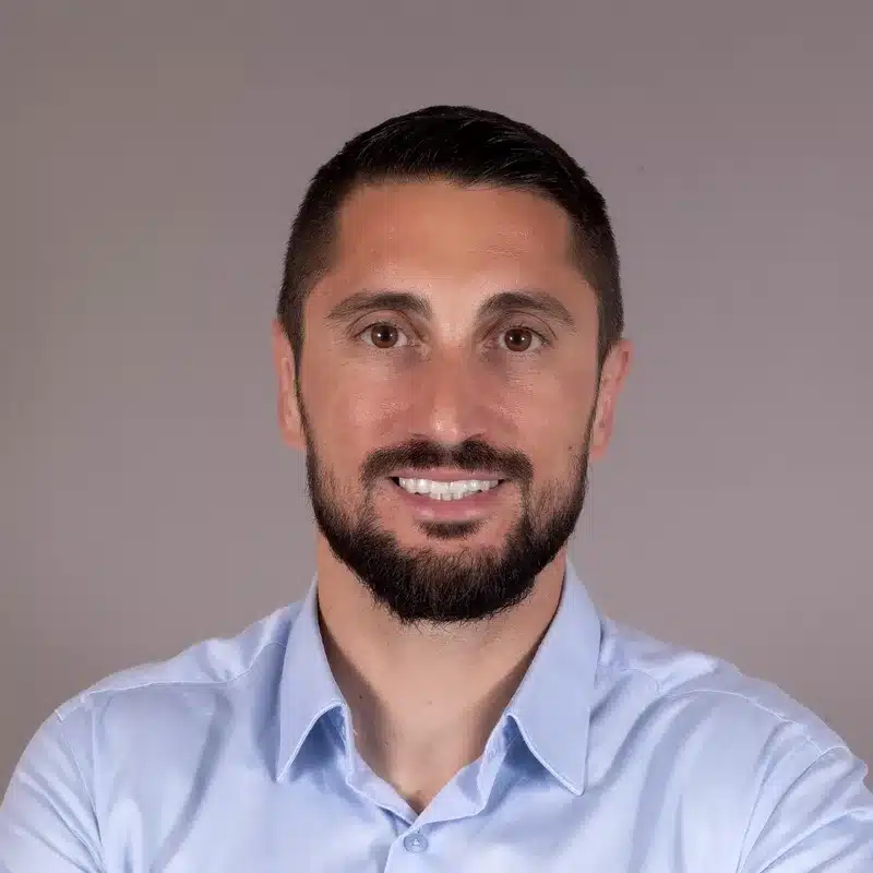 Homme souriant, cheveux bruns courts et barbe, chemise bleu clair, portrait professionnel pour un profil LinkedIn.