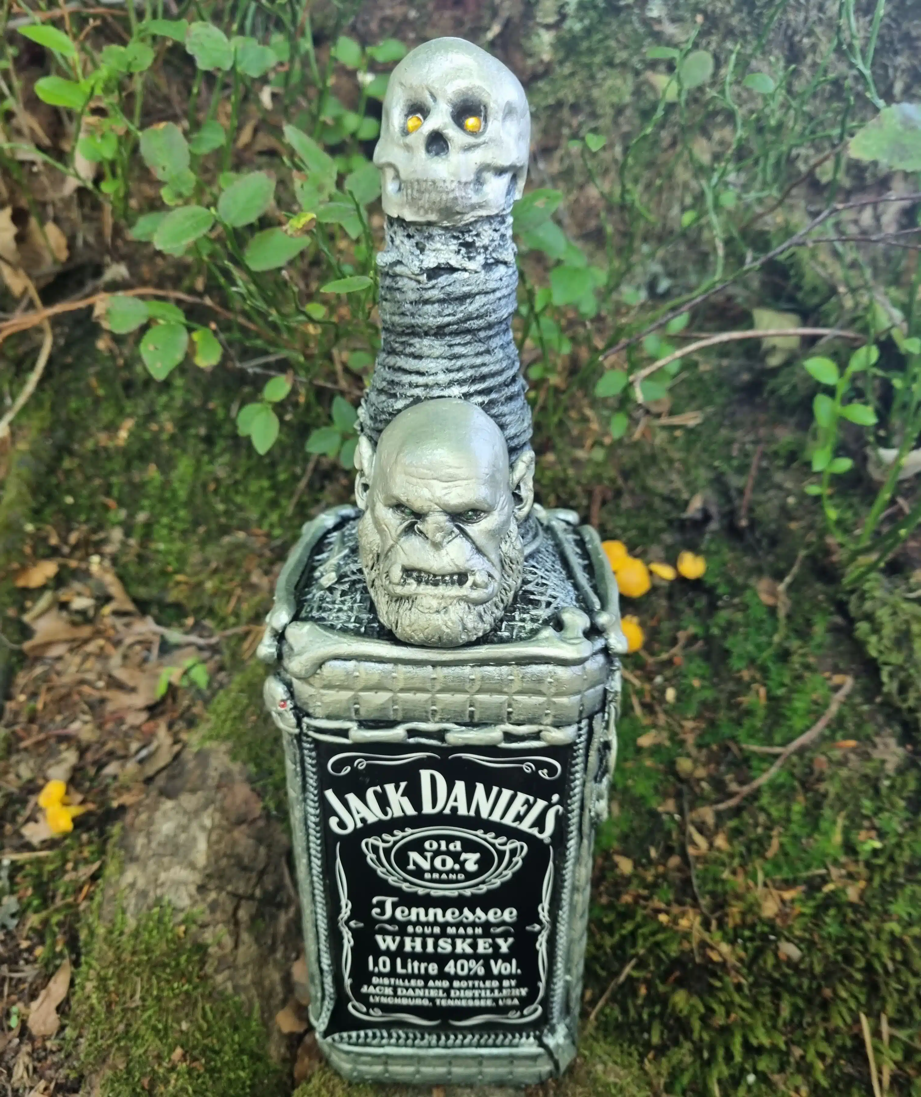 Bouteille décorative gothique – Créature & Crâne lumineux – Pièce unique 1 Une bouteille de Jack Daniels décorée de têtes de mort, posée à l'extérieur sur des plantes et de la mousse.