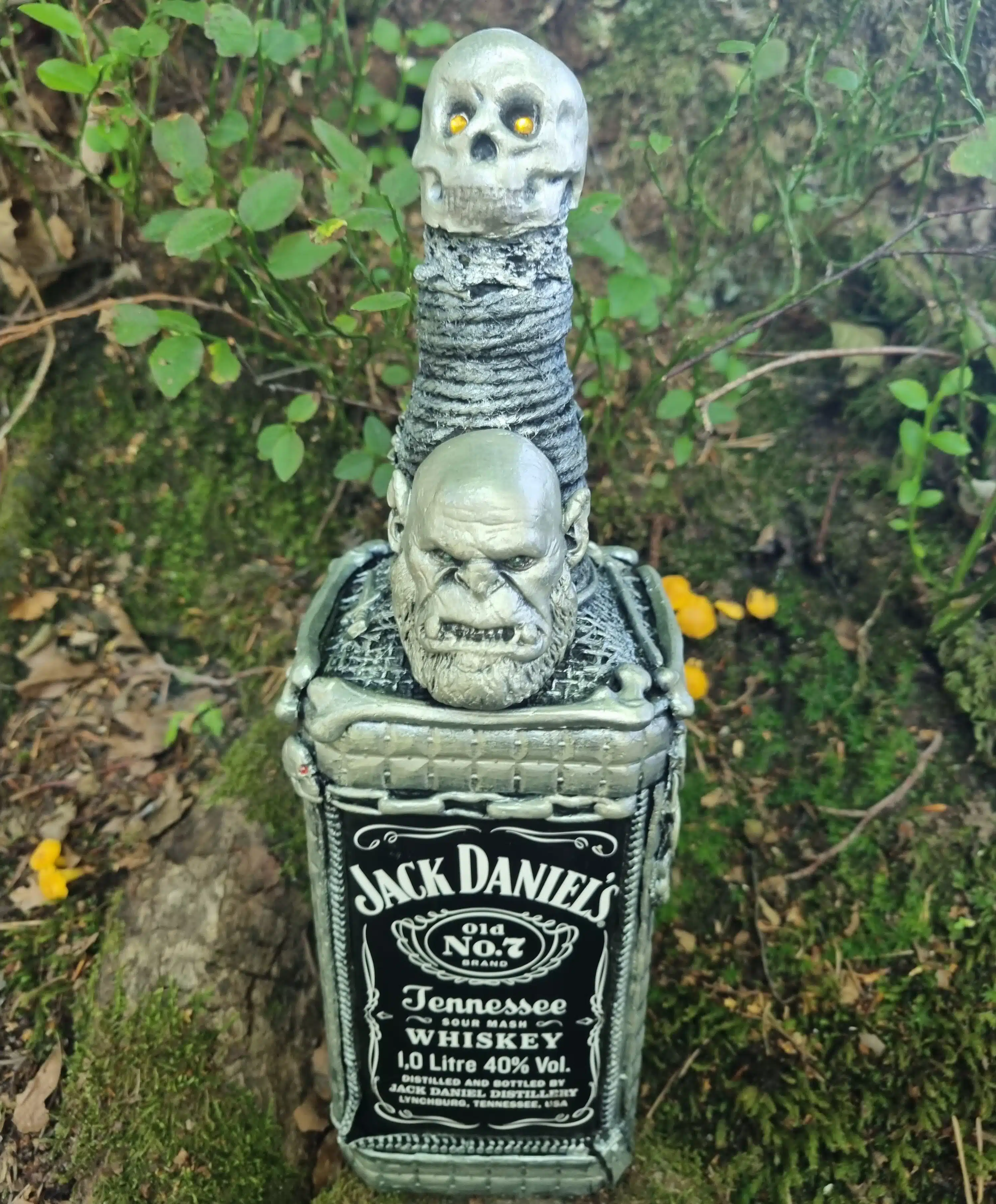 Bouteille décorative gothique – Créature & Crâne lumineux – Pièce unique 2 Une bouteille de Jack Daniels ornée de crânes et de têtes de zombies, posée au milieu de mousse, de plantes et d'un décor naturel en extérieur.