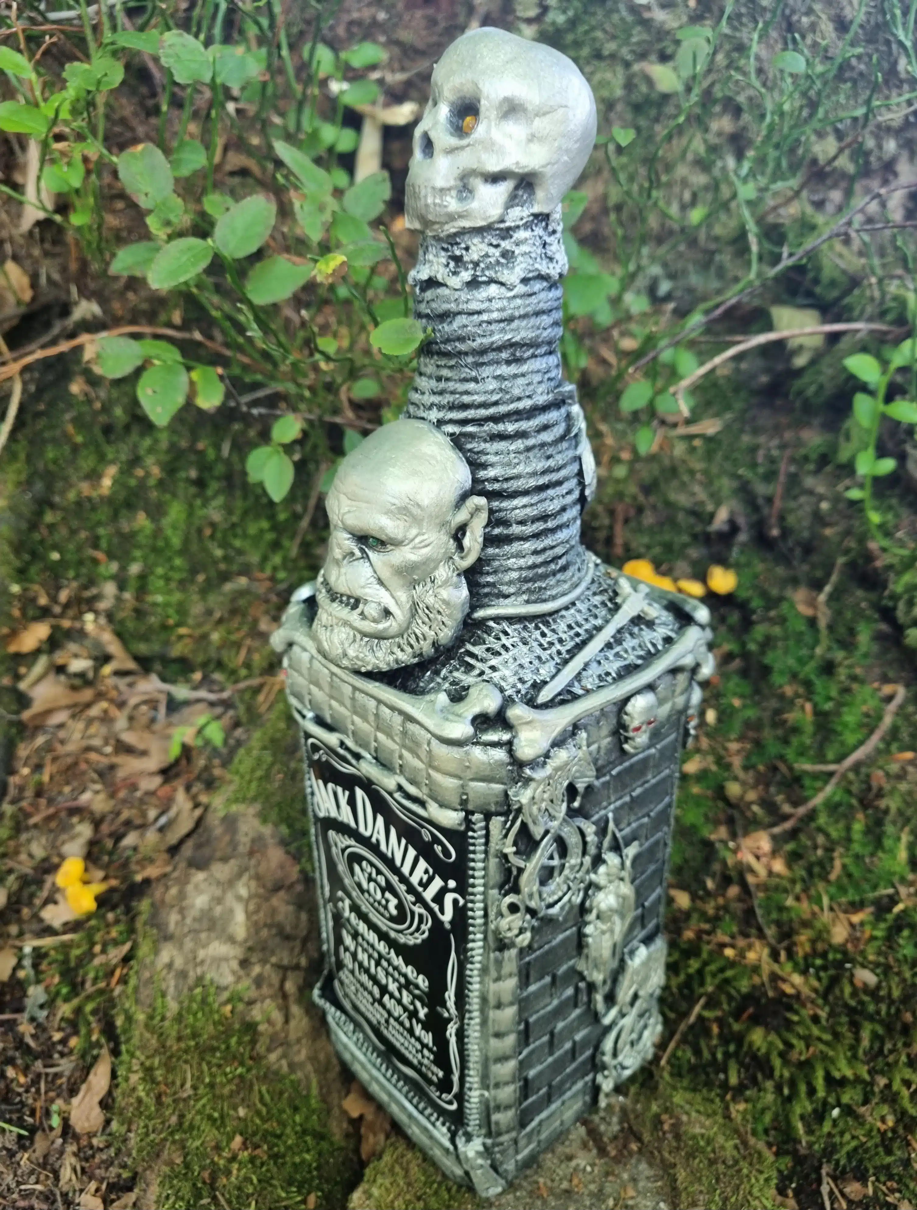 Bouteille décorative gothique – Créature & Crâne lumineux – Pièce unique 3 Bouteille de Jack Daniels décorée de crânes argentés et d’une tête d’orc sur un sol forestier moussu.