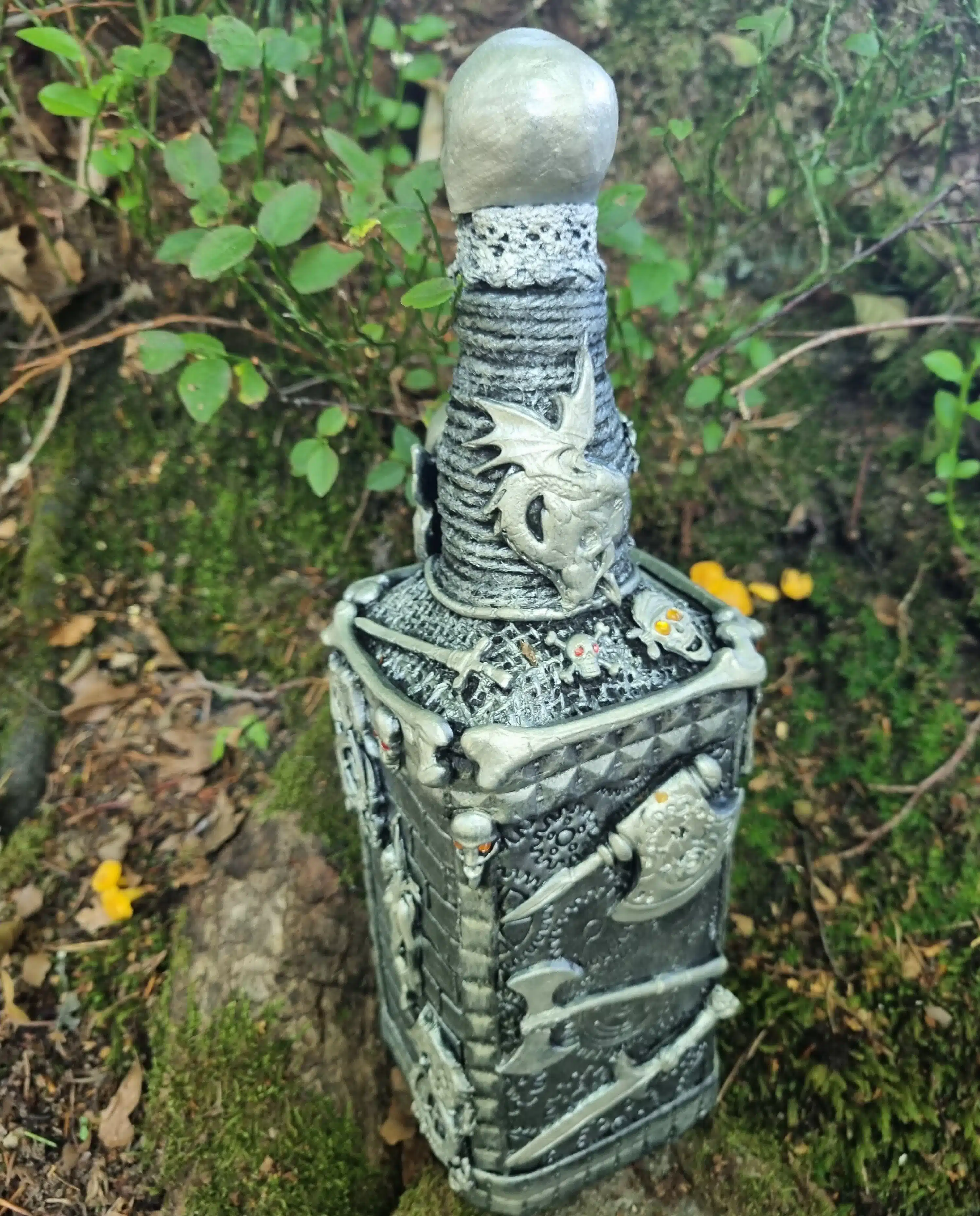 Bouteille décorative gothique – Créature & Crâne lumineux – Pièce unique 4 Bouteille décorative argentée ornée de dragons, de crânes, de haches et d'épées, posée sur un sol moussu à l'extérieur.