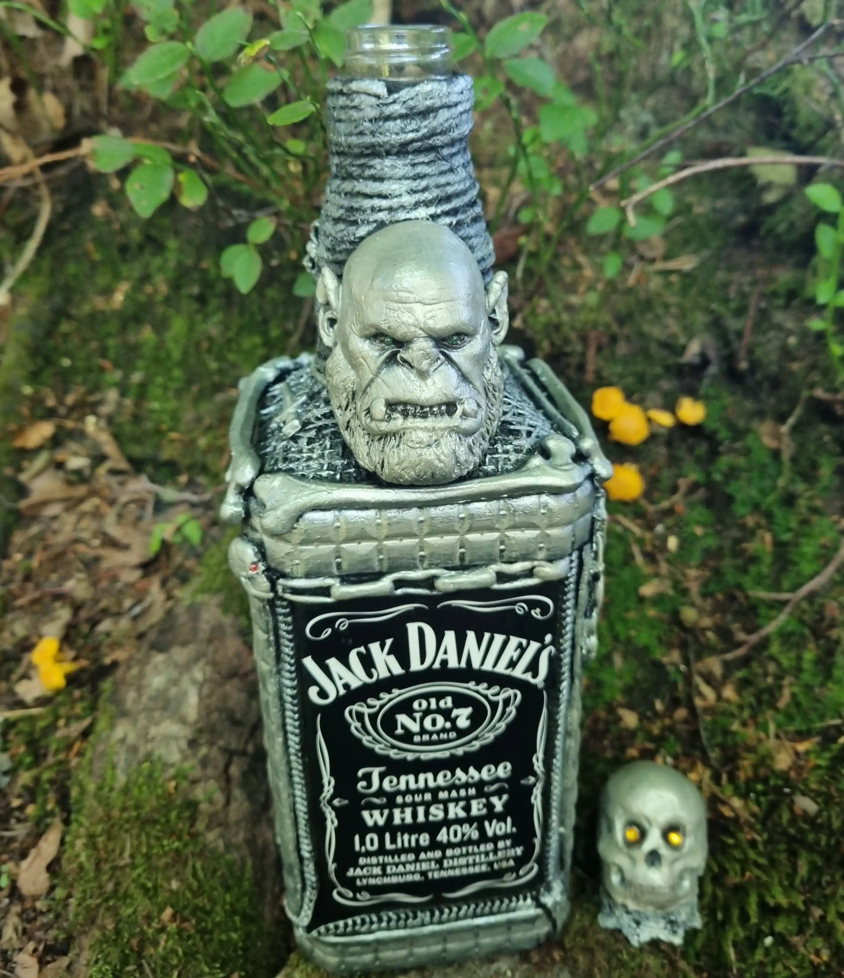 Bouteille décorative gothique – Créature & Crâne lumineux – Pièce unique 6 Une bouteille de Jack Daniels ornée d'une tête d'orc sculptée et d'un petit crâne, posée à l'extérieur près de plantes vertes.