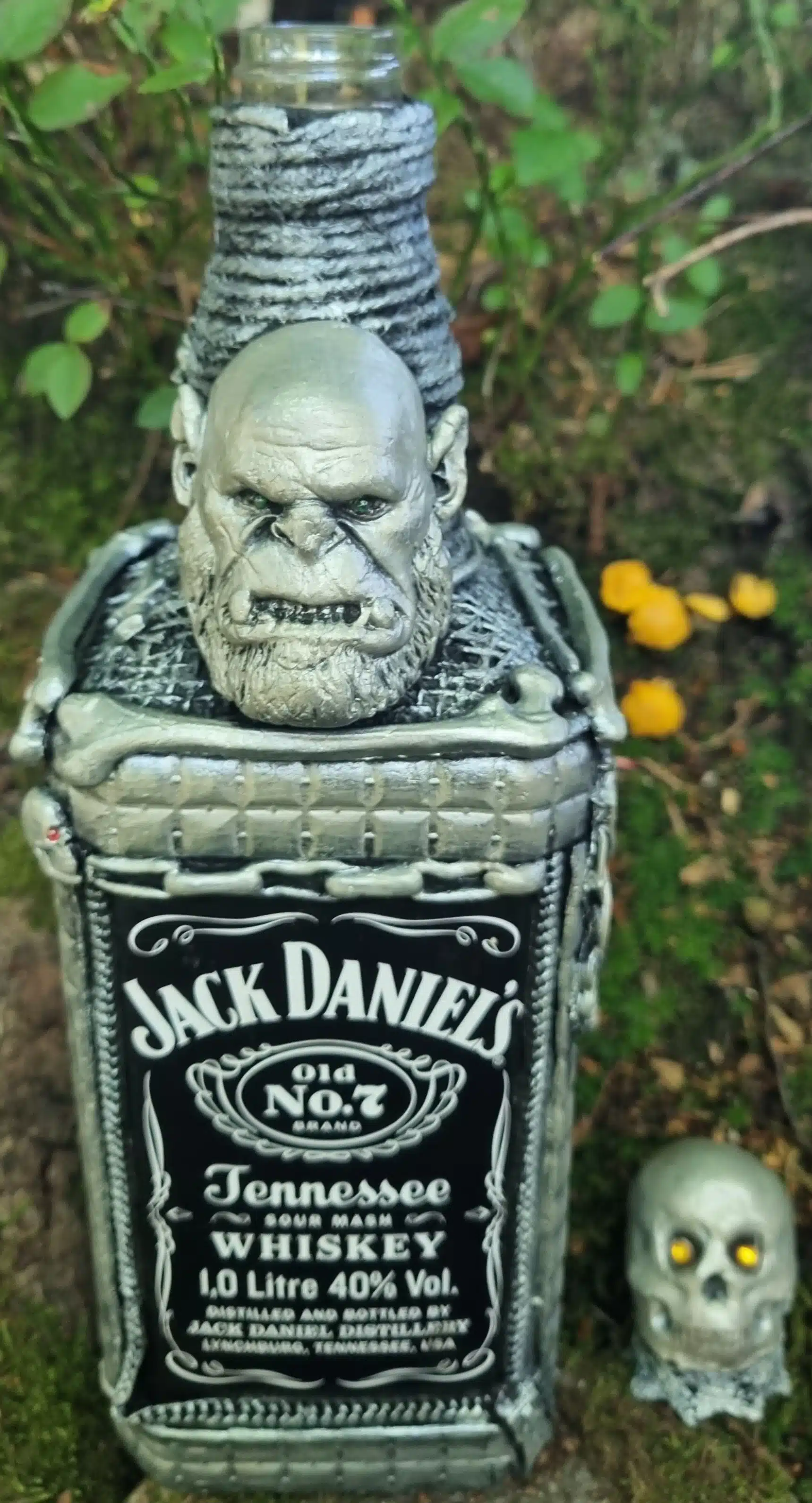 Bouteille décorative gothique – Créature & Crâne lumineux – Pièce unique 7 Bouteille de Jack Daniels ornée d’une tête d’ogre grise, d’une armure et d’un petit crâne posé sur de la mousse.