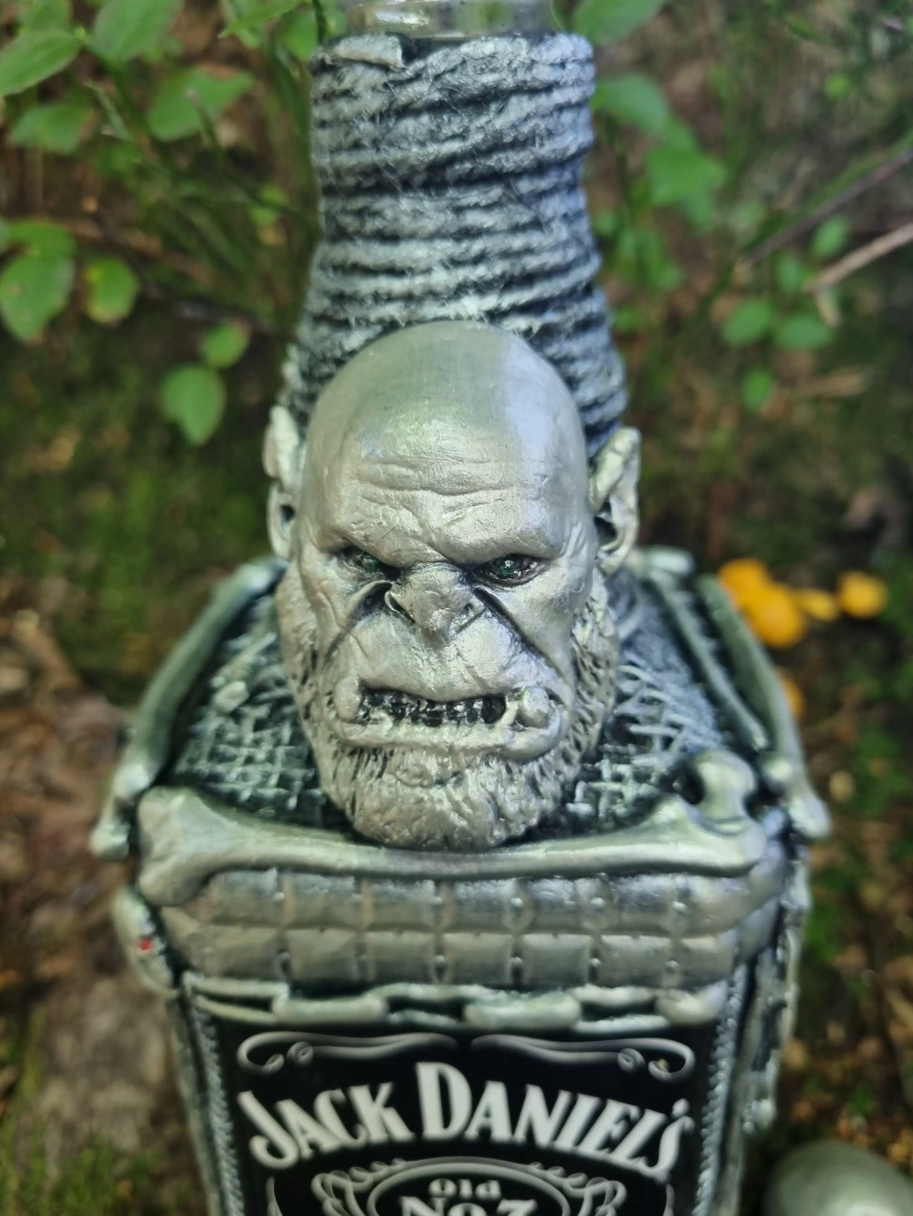 Bouteille décorative gothique – Créature & Crâne lumineux – Pièce unique 8 Une bouteille de Jack Daniels surmontée d’un visage monstrueux argenté, posée à l’extérieur au milieu des plantes.