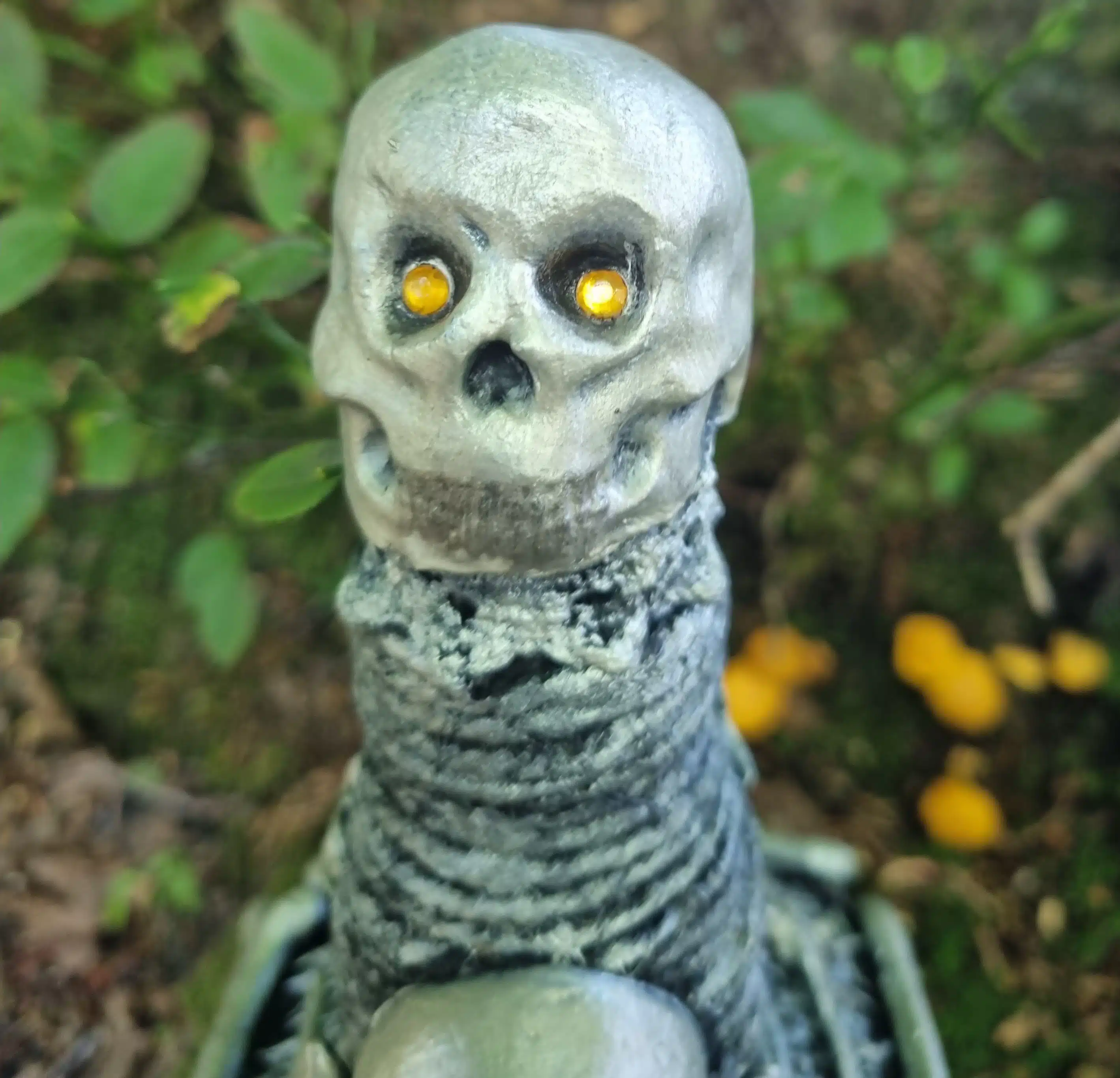 Bouteille décorative gothique – Créature & Crâne lumineux – Pièce unique 9 Statue squelettique grise aux yeux jaunes, entourée de plantes et de champignons jaunes en pleine nature.