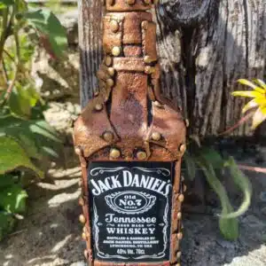 Accueil 12 Bouteille de Jack Daniels ornée de clous décoratifs, posée sur une pierre, avec des plantes en arrière-plan.