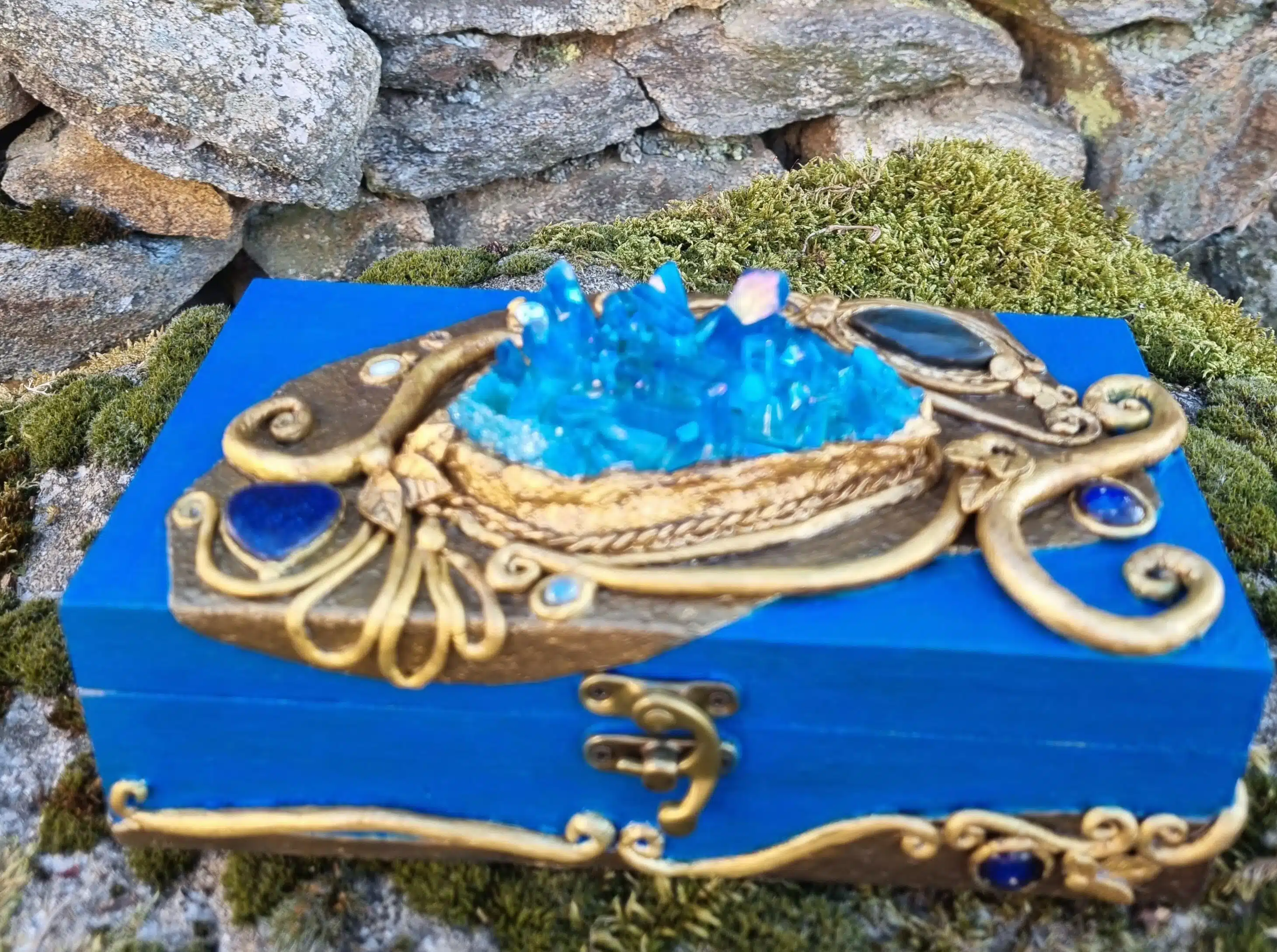 Coffret décoratif bleu & or – Cristaux – Pièce unique artisanale 3 Boîte bleue ornée de cristaux et de dorures, posée sur de la mousse devant un mur en pierre.