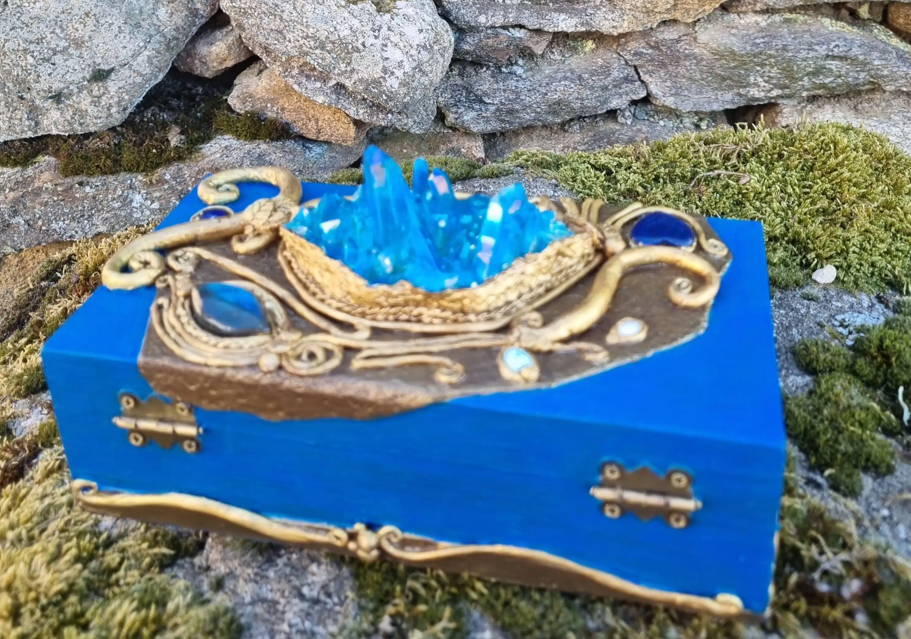 Coffret décoratif bleu & or – Cristaux – Pièce unique artisanale 6 Boîte en bois bleue ornée de motifs dorés et de cristaux, posée sur de la mousse devant un mur de pierre.
