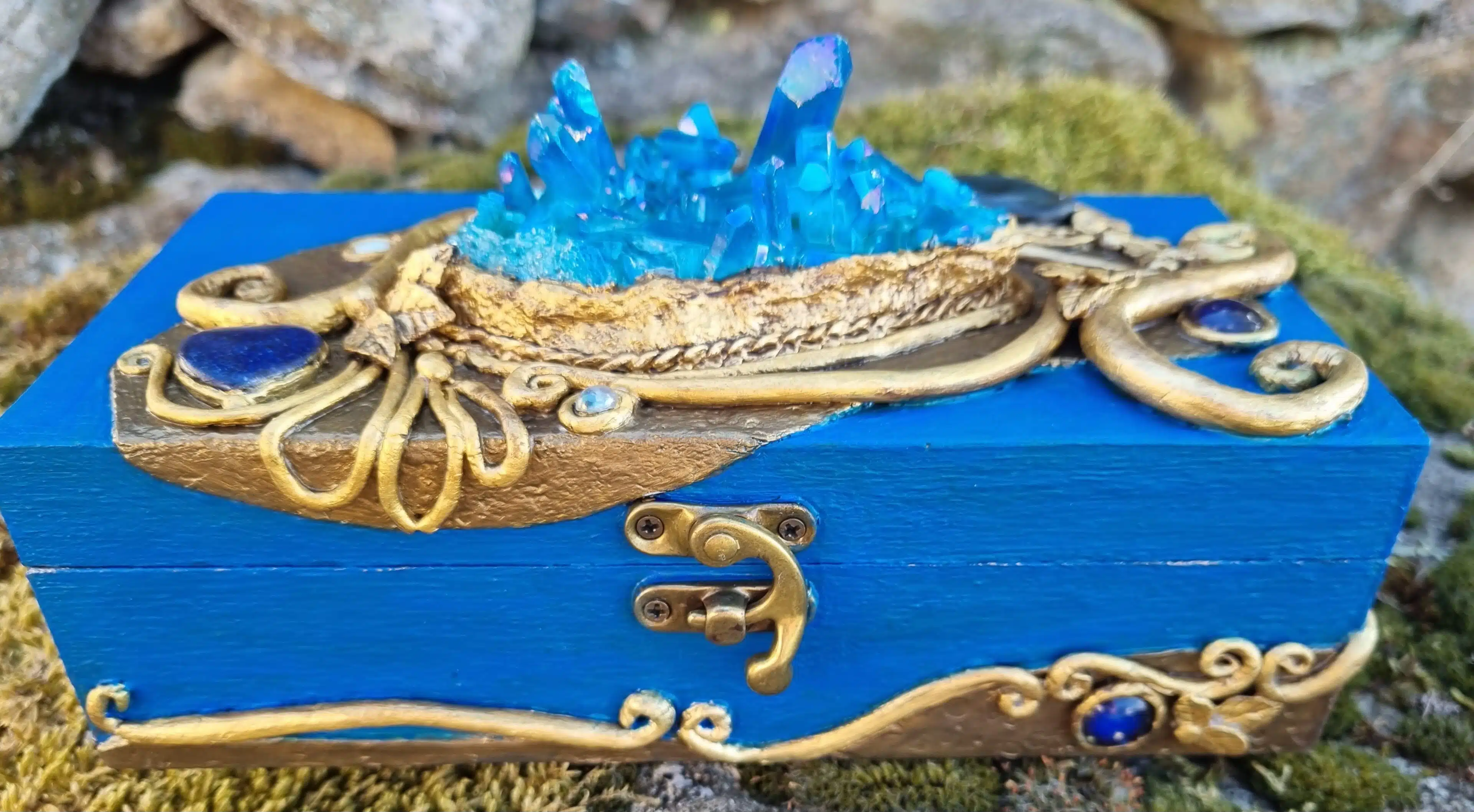 Coffret décoratif bleu & or – Cristaux – Pièce unique artisanale 11 Boîte en bois bleue ornée de cristaux et de dorures, posée sur de la mousse, avec des pierres en arrière-plan.