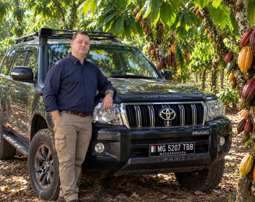 Un homme adossé à un Toyota Land Cruiser noir garé dans une plantation de cacao.